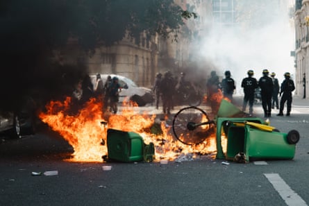 A line of riot police stand in a street behind overturned green wheelie bins and flames. Smoke is rising from the fire.