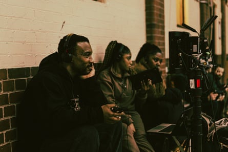 He checks a monitor with headphones on, with his team sitting on a bench around him