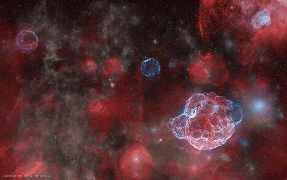 Imagem de longa exposição revela, em cores, a vasta constelação do Cocheiro. Nuvens de hidrogênio (vermelho), oxigênio (azul) e poeira abrigam remanescentes de supernovas, verdadeiros "fantasmas cósmicos" de estrelas que explodiram há milênios.